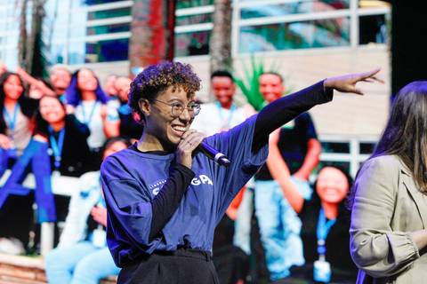 Apresentadora do Evento GPA no Espaço Unimed em São Paulo, apontando e interagindo com o público, convidando a participação e mostrando entusiasmo.'