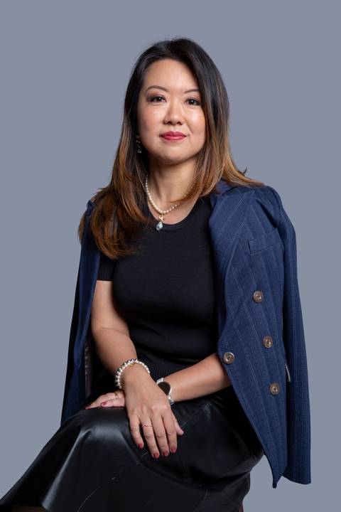 Mulher oriental de meia-idade, sentada em um ensaio corporativo em São Paulo, com um blazer azul marinho listrado casualmente solto sobre os ombros e blusa branca, mantendo um olhar sério e concentrado.'