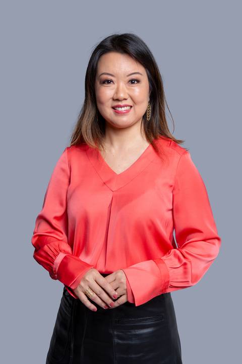 Mulher oriental de meia-idade, em pé para um ensaio corporativo em São Paulo, vestindo uma blusa salmão com um leve brilho, com as mãos unidas suavemente à frente, transmitindo uma postura elegante.'