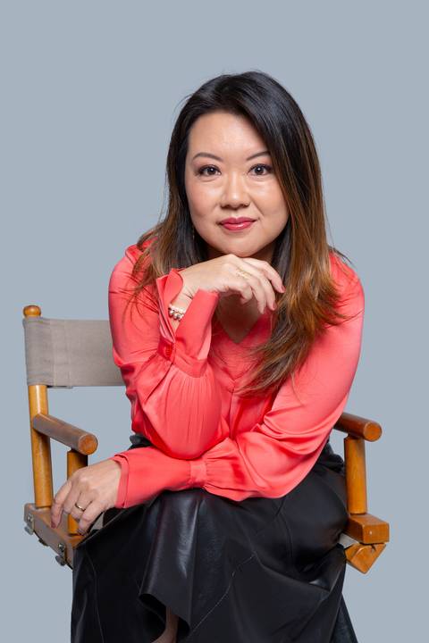 Mulher oriental de meia-idade, sentada em ensaio corporativo em São Paulo, com uma blusa salmão brilhante, o queixo delicadamente apoiado em uma das mãos, em um gesto pensativo e profissional.'