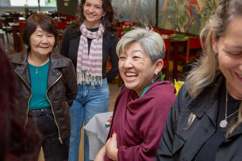 A aniversariante de 50 anos sorrindo radiantemente, cercada por convidados que compartilham a alegria da celebração no Restaurante Nanako em São Paulo.'