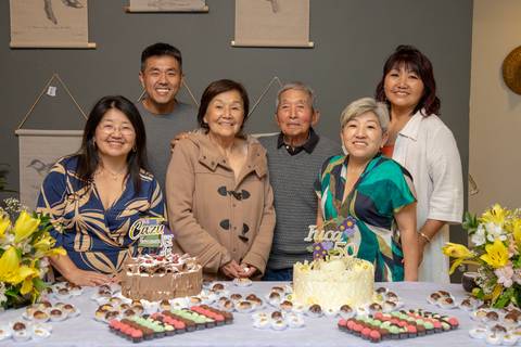 Foto de família dos aniversariantes, a mulher de 50 e o homem de 41 anos, com seus entes queridos, reunidos na mesa do bolo no Restaurante Nanako em São Paulo, celebrando a união.'