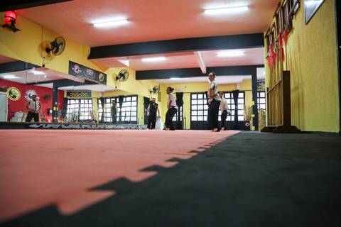Plano baixo do chão da Academia Kwan Kun com os participantes ao fundo durante o Exame de Faixa Preta. A imagem destaca a perspectiva do tatame e o ambiente de concentração marcial.'