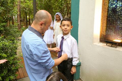 Pai auxiliando e arrumando seu filho, vestindo roupas formais para a Celebração Catequética na Paróquia Universitária Beato Carlo Acutis em São Paulo, reforçando a importância da participação familiar na vida religiosa.'