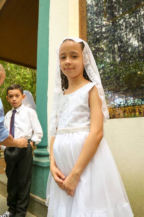 Criança menina vestindo a veste branca tradicional da catequese, sorrindo, em preparação para a Celebração Catequética na Paróquia Universitária Beato Carlo Acutis em São Paulo, simbolizando pureza e o rito religioso.
'