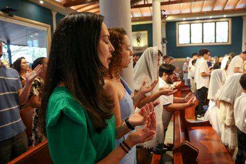 Fiéis em profunda oração durante a Celebração Catequética na Paróquia Universitária Beato Carlo Acutis, em São Paulo, com olhos fechados e mãos abertas em gesto de entrega e fé. A imagem transmite um instante de intimidade espiritual, conexão com Deus e d'