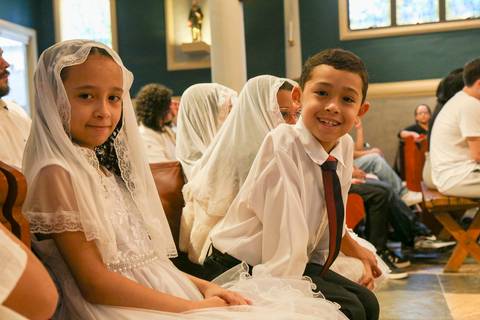 Grupo de crianças catequizandas, vestidas com suas vestes, olhando atentas para a câmera enquanto aguardam o início da Celebração Catequética na Paróquia Universitária Beato Carlo Acutis em São Paulo, transmitindo expectativa e inocência.'