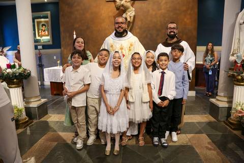 Crianças posando ao lado do padre ao final da Celebração Catequética na Paróquia Universitária Beato Carlo Acutis, em São Paulo. A fotografia registra o encerramento festivo da liturgia, com sorrisos, gratidão e o vínculo afetivo entre pastor e catequizan'