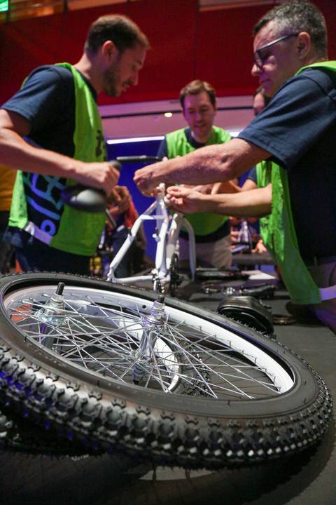 Close-up da roda de bicicleta em primeiro plano, com mãos de participantes da Plurix no Teatro B32 em São Paulo trabalhando em equipe na montagem, durante atividade corporativa'