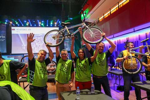 Participantes da Plurix comemorando com entusiasmo a montagem bem-sucedida de uma bicicleta, em atividade de trabalho em equipe no Teatro B32 em São Paulo'
