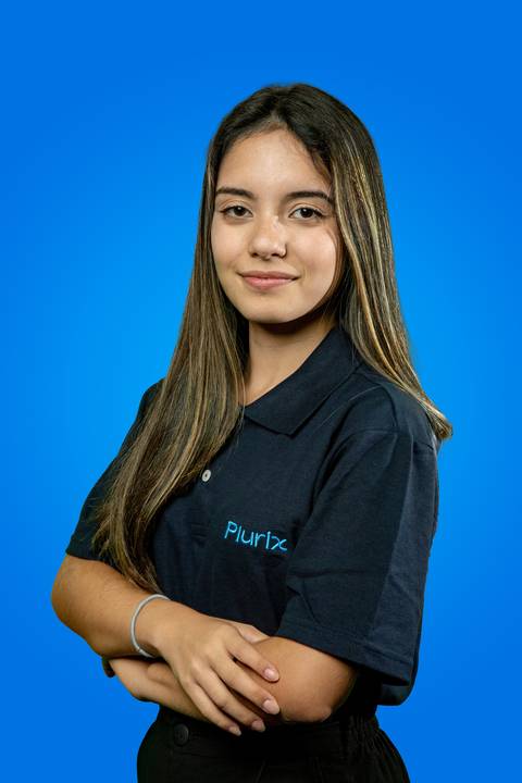 Retrato profissional de moça com sorriso discreto (sem mostrar os dentes) e braços cruzados, vestindo camisa polo da Plurix em frente a um fundo azul corporativo'