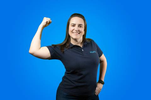Retrato profissional de mulher sorridente vestindo camisa polo da Plurix, fazendo o gesto de 'força feminina' com o punho cerrado, em frente a um fundo azul corporativo'