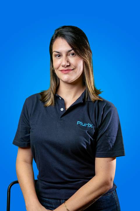 Retrato profissional de mulher sentada e sorrindo, vestindo camisa polo da Plurix em frente a um fundo azul corporativo'
