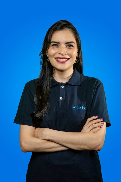 Retrato profissional de mulher sorrindo de frente, com braços cruzados e vestindo camisa polo da Plurix, em frente a um fundo azul corporativo'