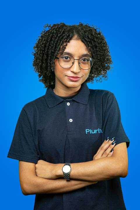 Retrato profissional de mulher jovem com cabelo cacheado, expressão séria e confiante, usando óculos e com braços cruzados, vestindo camisa polo da Plurix em frente a um fundo azul corporativo'