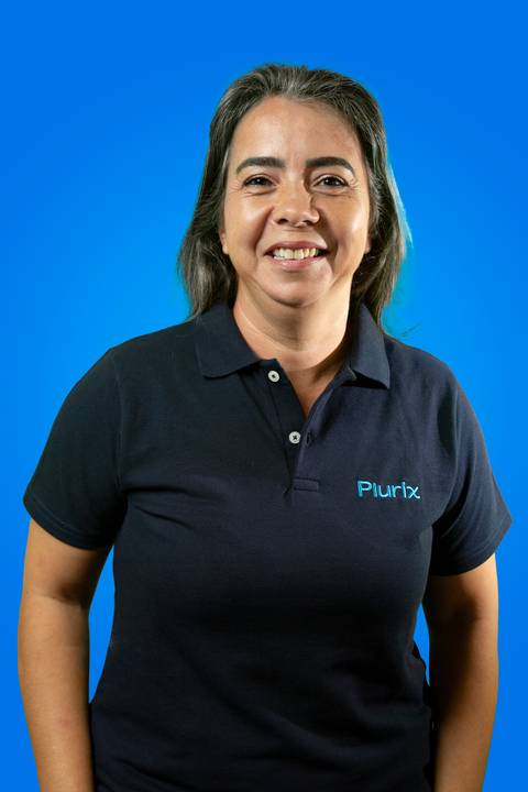 Retrato profissional de senhora sorridente vestindo camisa polo da Plurix, em frente a um fundo azul corporativo que remete à identidade visual da empresa'