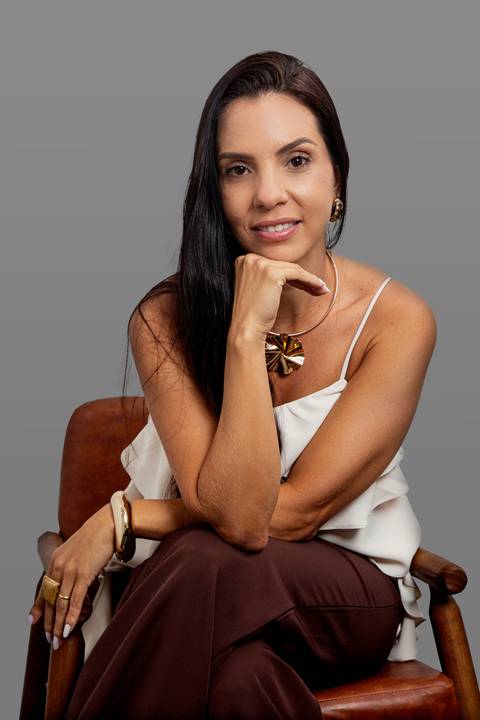 Retrato corporativo de mulher de negócios sentada, encarando a câmera com expressão confiante e queixo apoiado na mão, vestindo blusa branca, em fundo cinza profissional'