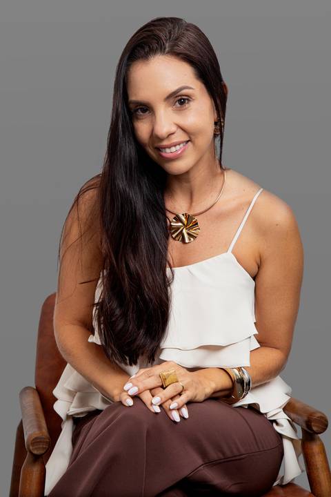 Retrato corporativo de mulher de negócios sentada, olhando diretamente para a câmera com uma expressão profissional e acolhedora, vestindo blusa branca, em fundo cinza'