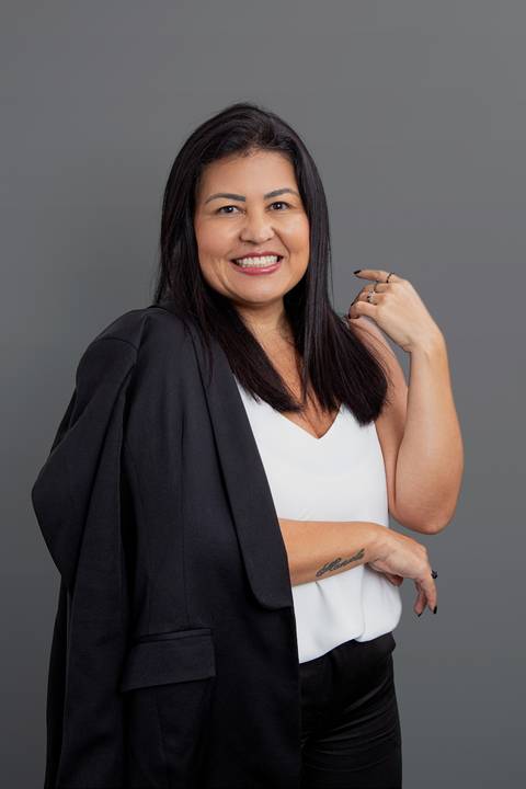 Retrato corporativo de mulher de negócios com terno preto elegantemente apoiado em um ombro e camisa branca, posando com uma mão na cintura e a outra levantada, em fundo cinza profissional'
