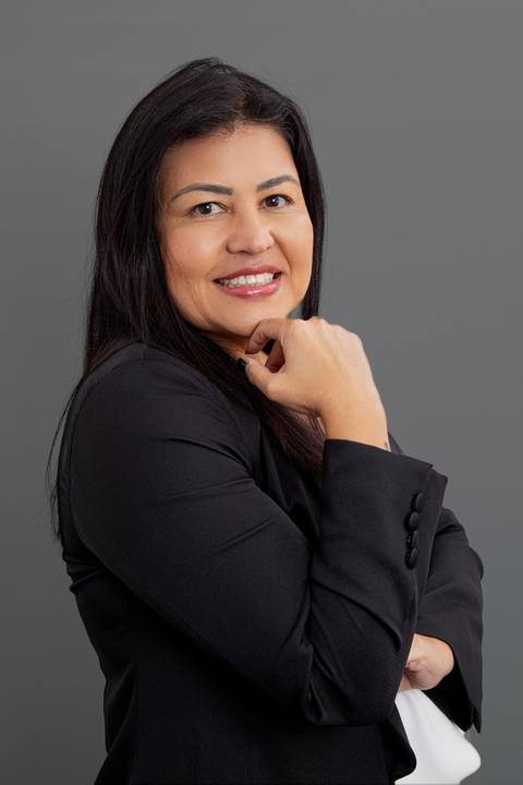 Retrato corporativo de mulher de negócios com terno preto levemente de lado e camisa branca, posando com a mão da frente elegantemente levantada na altura do queixo, em fundo cinza profissional'