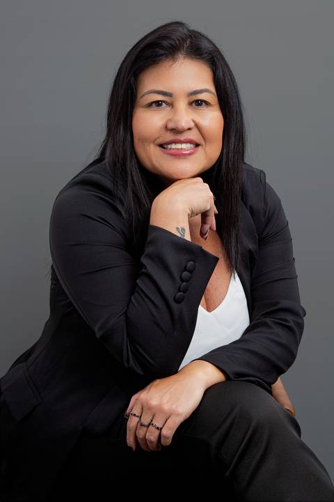 Retrato corporativo de mulher de negócios sentada, encarando a câmera com expressão confiante e queixo apoiado na mão, vestindo terno preto e camisa branca, em fundo cinza profissional'