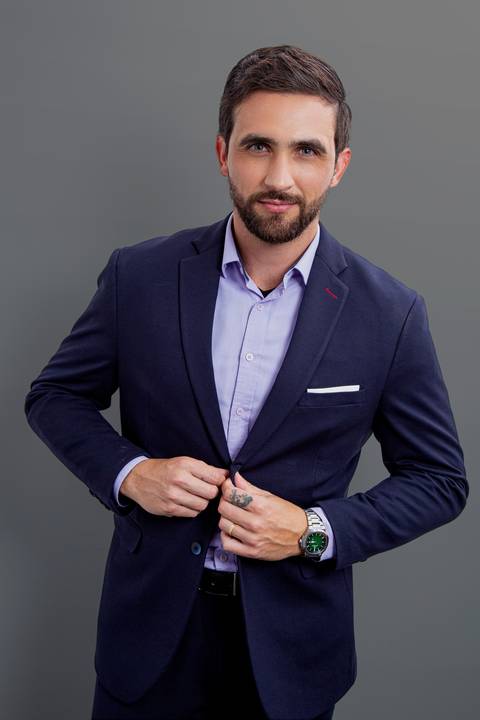 Retrato corporativo de homem de negócios de pé, levemente inclinado para frente com as mãos nos bolsos do terno azul marinho, em fundo cinza profissional'