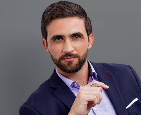 Close-up corporativo de homem de negócios com terno azul marinho e camisa azul (sem gravata), sentado levemente inclinado com a mão levantada e olhar sério, em fundo cinza profissional'