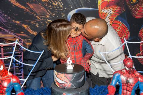 Momento afetuoso na festa de 6 anos do menino fantasiado de Homem-Aranha (sem máscara) no Kid Recanto Buffet em São Paulo: pais beijando o filho sorridente na bochecha, com o bolo e cenário do Homem-Aranha ao fundo'