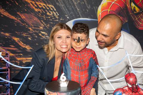 Foto em close do menino aniversariante fantasiado de Homem-Aranha (sem máscara) sorrindo entre seus pais, com o bolo e a decoração temática parcialmente visíveis ao fundo no Kid Recanto Buffet em São Paulo'