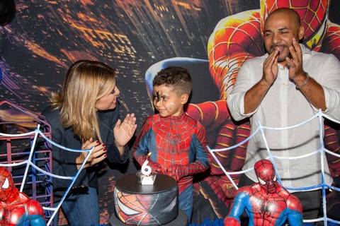 Momento do parabéns na festa de 6 anos do menino fantasiado de Homem-Aranha (sem máscara) no Kid Recanto Buffet em São Paulo, com o bolo iluminado e a família reunida'