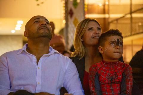 Família assistindo emocionada a um vídeo de fotos no Kid Recanto Buffet em São Paulo: pai e mãe com o filho fantasiado de Homem-Aranha (sem máscara) no colo, revivendo momentos especiais'