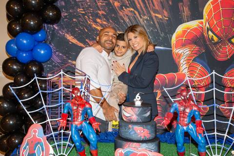 Menino aniversariante sorrindo ao posar com o pai e a mãe atrás do bolo decorado com tema do Homem-Aranha no Kid Recanto Buffet em São Paulo, celebrando seu aniversário de 6 anos'