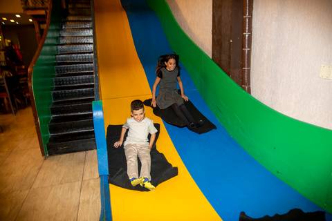 Menino aniversariante e menina descendo juntos e sorrindo por um escorregador colorido grande no Kid Recanto Buffet em São Paulo, em um momento de brincadeira compartilhada na festa de aniversário infantil'
