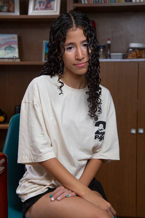 Retrato da filha sentada à mesa com pernas cruzadas, tendo ao fundo uma estante de madeira grande com livros e adornos em ensaio de família em SP'