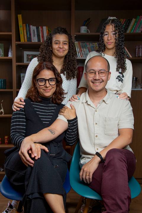 Retrato de família com pai e mãe sentados em cadeiras, filhas posicionadas atrás deles, e ao fundo uma grande estante de madeira, transmitindo união familiar em ensaio de família em SP'