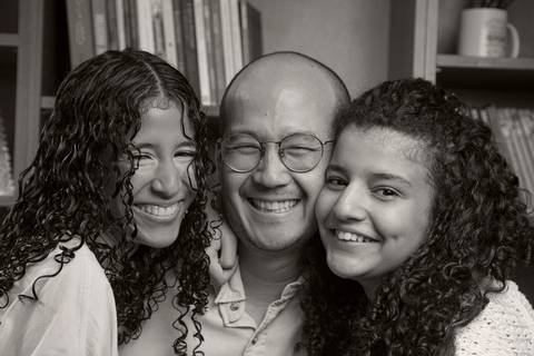 Foto em preto e branco de pai no centro com uma filha em cada lado, posando em frente a uma grande estante de madeira ao fundo, em retrato familiar em ensaio de família em SP'