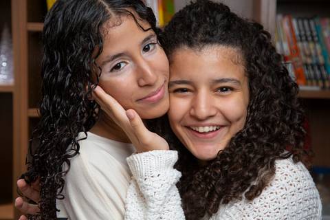 etrato das duas filhas, uma segurando o rosto da outra em gesto de carinho, posando em frente a uma grande estante de madeira ao fundo em ensaio de família em SP'