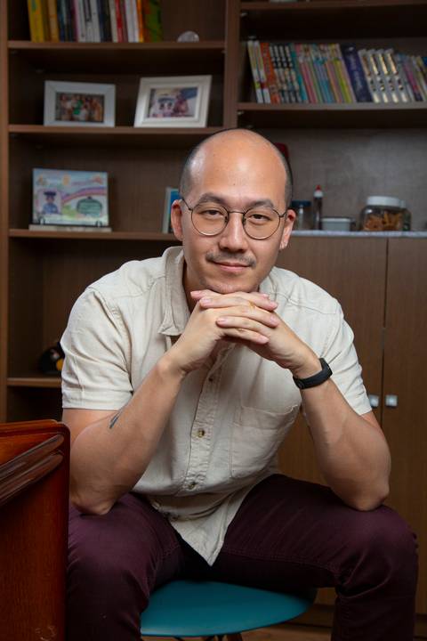 Retrato de pai sentado, com dedos entrelaçados e queixo apoiado nas mãos, cotovelos nos joelhos, em pose pensativa, com estante de madeira ao fundo em ensaio de família em SP'