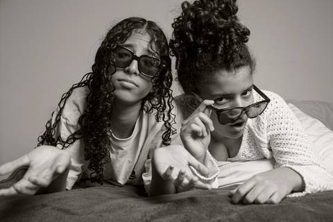 Foto em preto e branco das duas filhas na cama usando óculos escuros; uma posa com as mãos abertas, enquanto a outra segura os óculos e encara a câmera, em um retrato divertido em ensaio de família em SP'