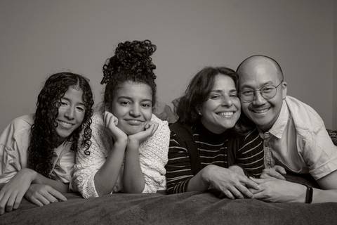 Foto em preto e branco de família deitada na cama, com as duas filhas, mãe e pai em sequência, em um retrato familiar íntimo e sereno em ensaio de família em SP'