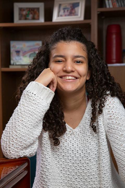 Retrato da filha sentada à mesa em pose relaxada, com o cotovelo apoiado e o rosto na mão, transmitindo tranquilidade em ensaio de família em SP'