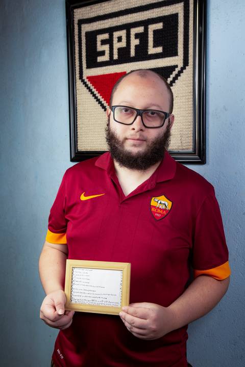 Filho segurando um acróstico emoldurado, presente feito por seu falecido pai, com parede azul e quadro do SPFC ao fundo, em um momento de memória e homenagem em ensaio de família em Mauá'