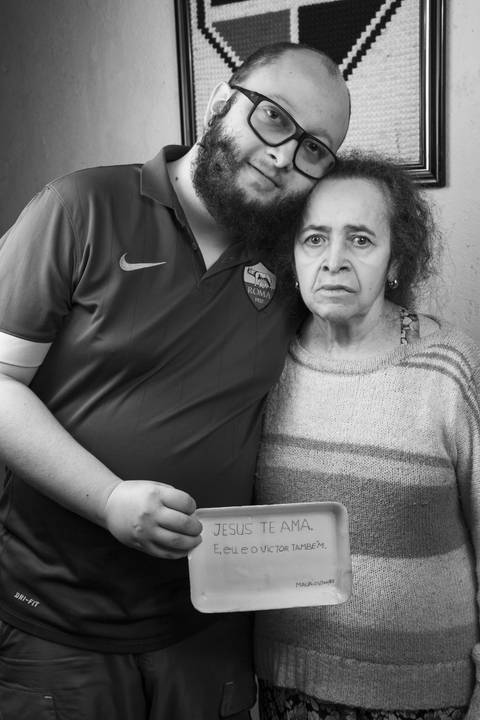 Foto em preto e branco de mãe e filho segurando um pratinho de isopor com mensagem de apoio escrita pelo falecido pai e filho para a mãe, em frente à parede azul com quadro do SPFC, expressando suporte familiar em ensaio de família em Mauá'