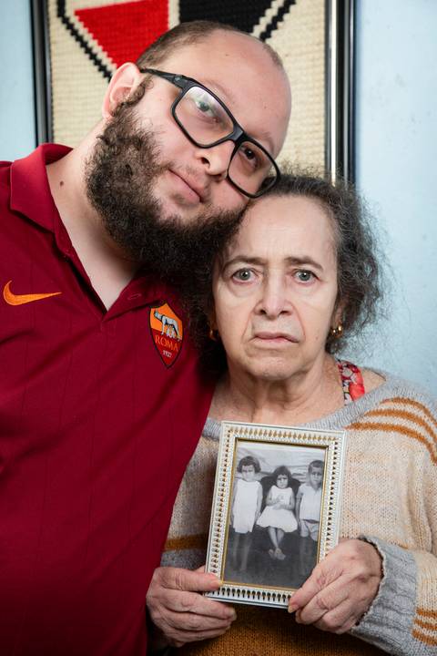 Retrato sério de mãe e filho posando em frente à parede azul com quadro do SPFC, segurando uma fotografia emoldurada da mãe com suas irmãs quando crianças em ensaio de família em Mauá'