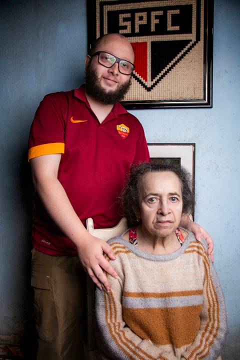 Retrato de mãe e filho com luz suave, posando juntos em frente a uma parede azul com um quadro decorativo e outro com o símbolo do SPFC em tecido em ensaio de família em Mauá'