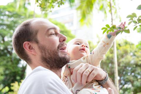 Pai segurando bebê com olhar de ternura, enquanto o bebê curioso toca um galho de árvore, em momento de descoberta na natureza em ensaio de família em SP'