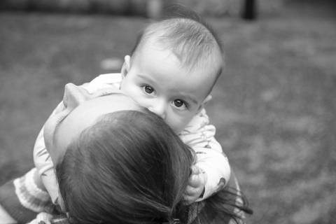 Foto em preto e branco de mãe de costas segurando bebê que olha diretamente para a câmera, em um momento de conexão em ensaio de família em SP'