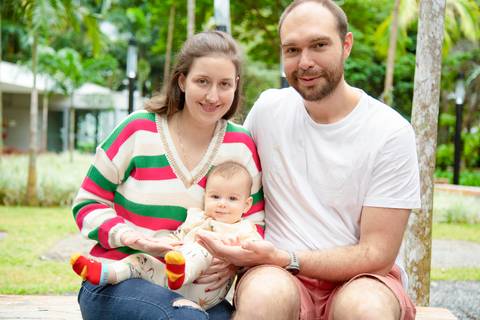 Retrato de família feliz com pai e mãe sentados em banco, bebê no colo da mãe, com fundo de vegetação verde exuberante em ensaio de família em SP'