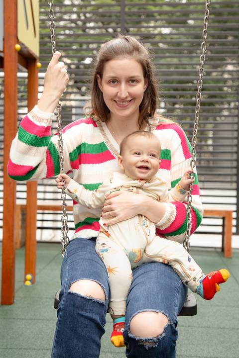Mãe sorrindo sentada em balanço de parque com bebê feliz no colo, em momento de diversão e carinho em ensaio de família em SP'