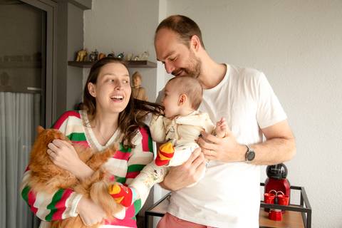 Momento espontâneo de família: mãe segurando gato laranja, pai segurando bebê que morde o cabelo da mãe, todos sorrindo em ensaio de família em SP'